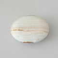 A polished oval stone or agate slab with white, beige, and brown banding, shown next to a clear plastic ruler on a light gray background. Available at J.W.H. Designs.