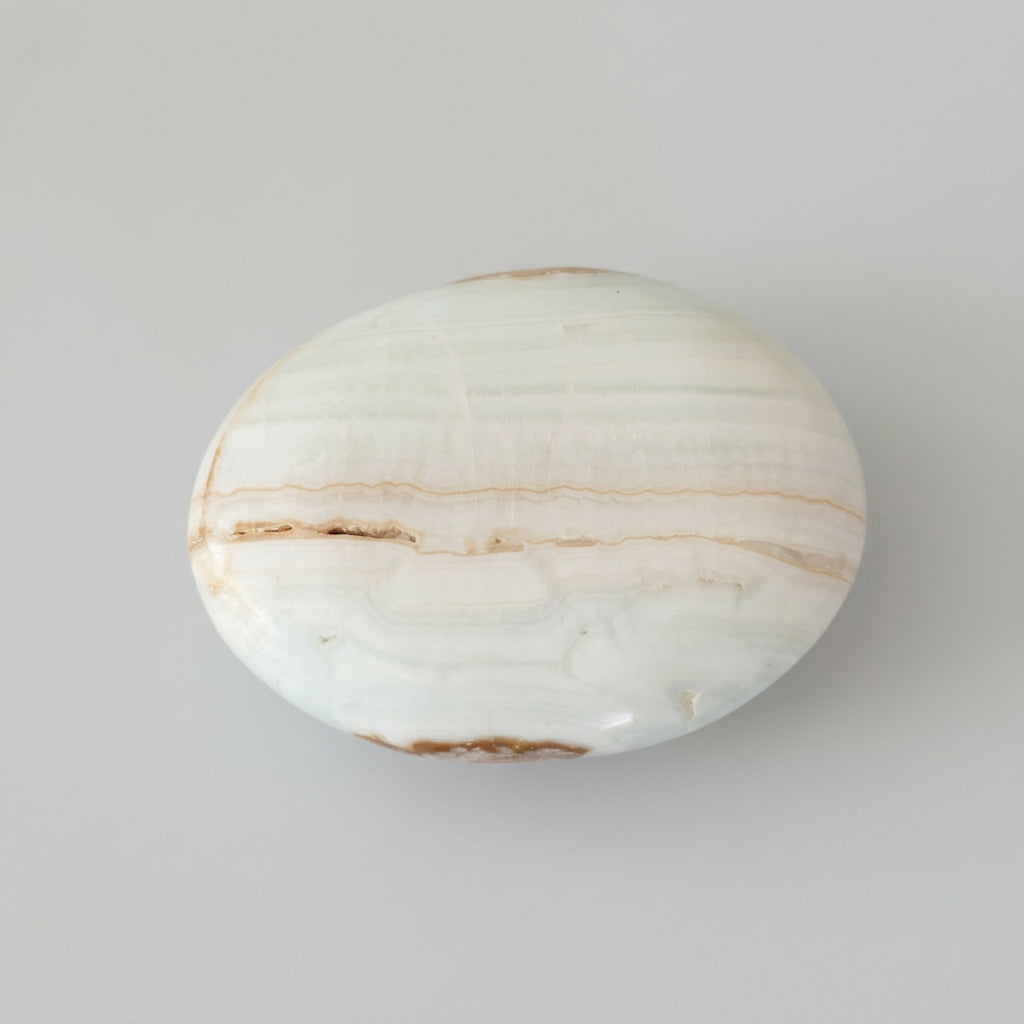 A polished oval stone or agate slab with white, beige, and brown banding, shown next to a clear plastic ruler on a light gray background. Available at J.W.H. Designs.