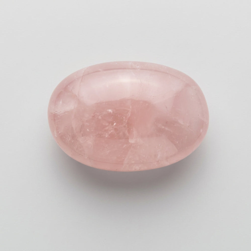A smooth, polished oval-shaped rose quartz crystal stone with a soft pink color, shown next to a clear plastic ruler for scale on a light gray background. Available at J.W.H. Designs, Canada.